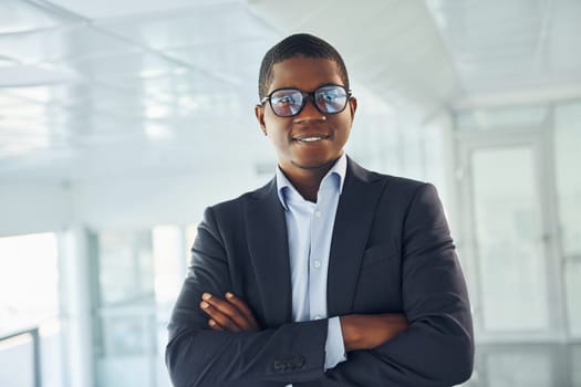 With arms crossed. Young african american businessman in black suit is indoors.