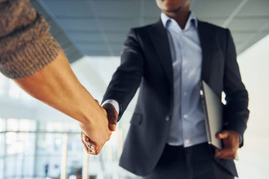 Doing handshake with customer. Young african american businessman in black suit is indoors.