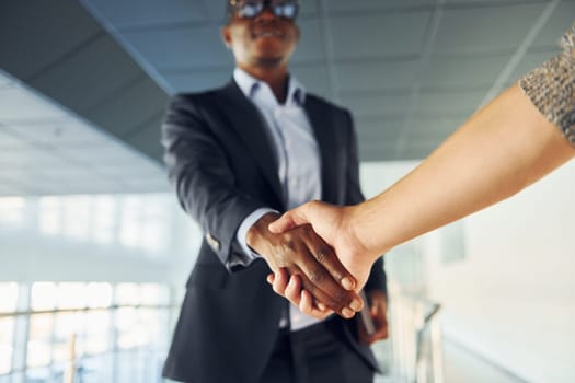 Doing handshake with customer. Young african american businessman in black suit is indoors.