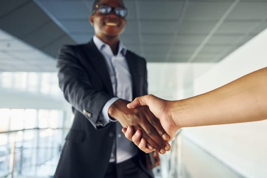 Doing handshake with customer. Young african american businessman in black suit is indoors.