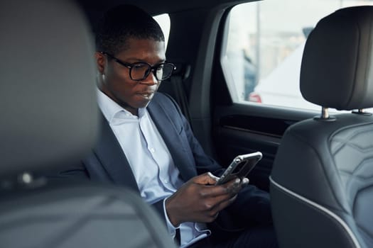 In glasses. Young african american businessman in black suit is in the automobile.