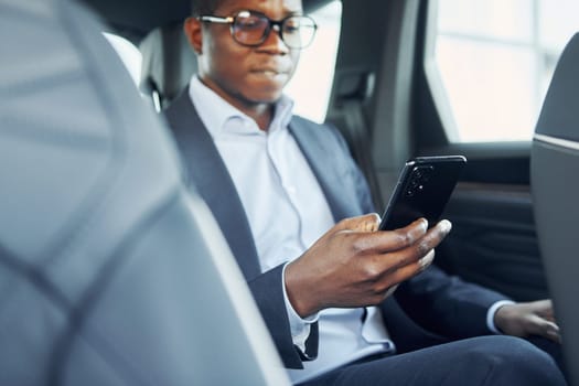 In glasses. Young african american businessman in black suit is in the automobile.