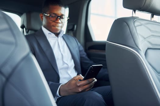 In glasses. Young african american businessman in black suit is in the automobile.
