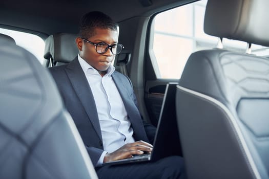 Passanger on the back seat. Young african american businessman in black suit is in the automobile.