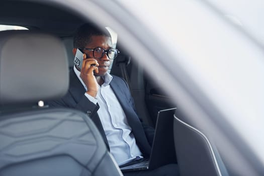 Passanger on the back seat. Young african american businessman in black suit is in the automobile.