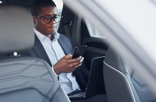Passanger on the back seat. Young african american businessman in black suit is in the automobile.