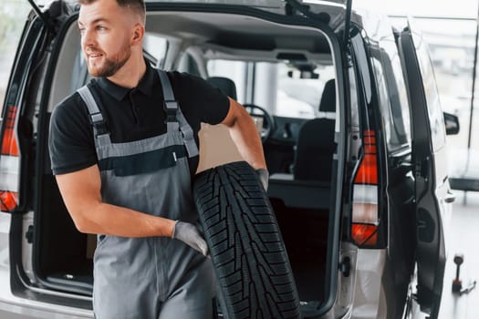 Holding tire. Man in uniform is working in the autosalon at daytime.