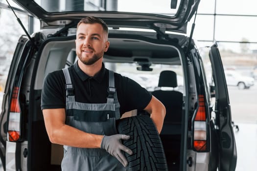 Holding tire. Man in uniform is working in the autosalon at daytime.