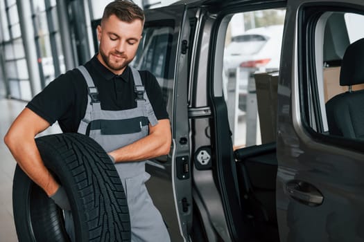 With tire in hands. Man in uniform is working in the autosalon at daytime.