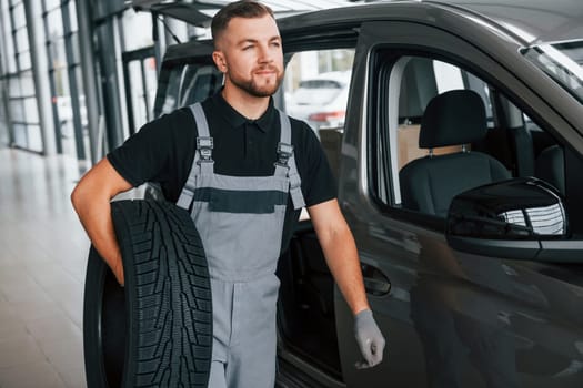 With tire in hands. Man in uniform is working in the autosalon at daytime.