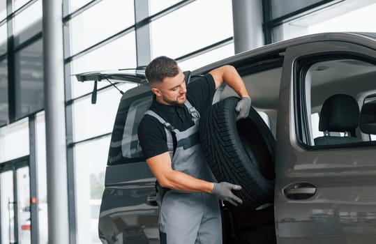 With tire in hands. Man in uniform is working in the autosalon at daytime.