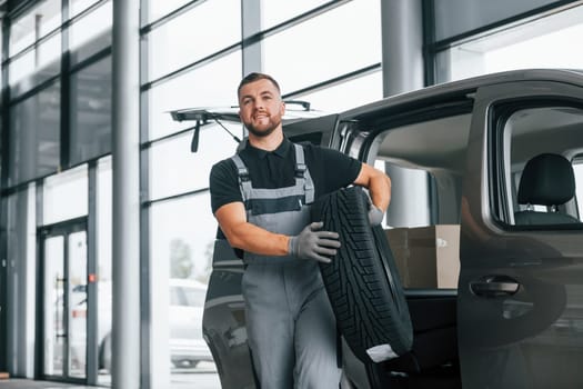 With tire in hands. Man in uniform is working in the autosalon at daytime.