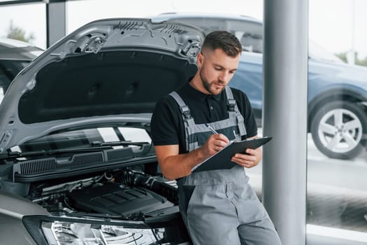 With notepad. Man in uniform is working in the autosalon at daytime.