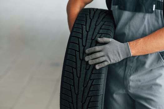 Man in uniform is working in the autosalon at daytime. Holding tire in hands.