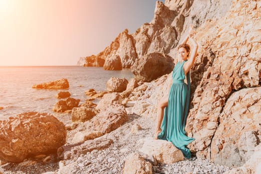 Side view a Young beautiful sensual woman in a red long dress posing on a rock high above the sea during sunrise. Girl on the nature on blue sky background. Fashion photo.