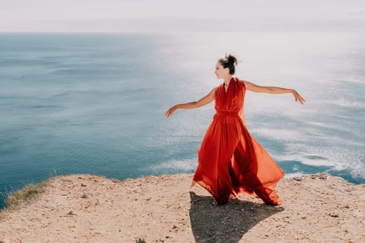 Side view a Young beautiful sensual woman in a red long dress posing on a rock high above the sea during sunrise. Girl on the nature on blue sky background. Fashion photo.