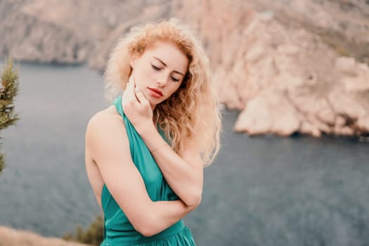 Side view a Young beautiful sensual woman in a mint long dress posing on a volcanic rock high above the sea during sunset. Girl on the nature on overcast sky background. Fashion photo