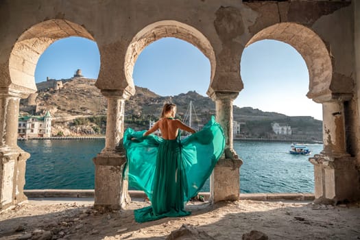 Rear view of a happy blonde woman in a long mint dress posing against the backdrop of the sea in an old building with columns. Girl in nature against the blue sky