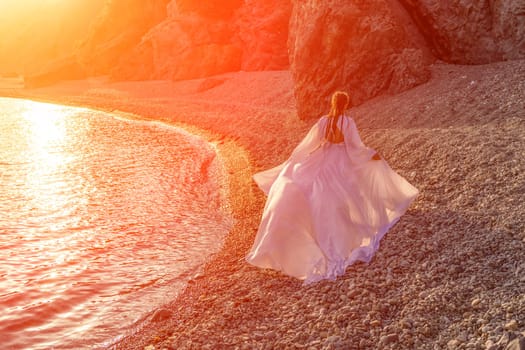 Mysterious woman silhouette long hair walks on the beach ocean water, sea nymph wind listens to the wave. Throws up a long white dress, a divine sunset. Artistic photo from the back without a face.