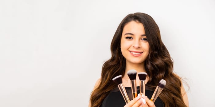 Positive attractive woman holding cosmetic makeup brushes over gray background with copy space.