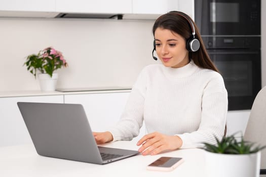 Female leader in headset discusses details of project with team via Notebook. Young brunette woman sits at table in kitchen working online slow motion