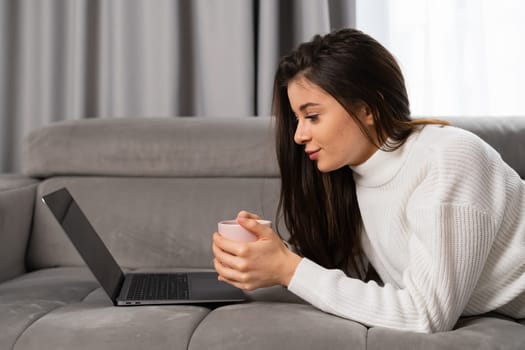 Young beautiful woman relaxing, holding a cup, watching movie using a laptop, laying on the sofa