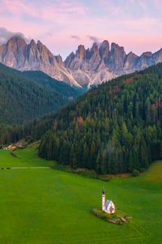 The fantastic landscape of Santa Maddalena village with St. Johann old church and Dolomites Geisler mountain group in valley Val di Funes at pink sunset.