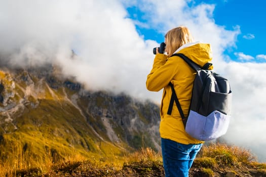 Young blonde photographer in yellow sport jacket takes pictures of Passo Giau pass using camera. Woman with backpack enjoys activity in Italian Alps
