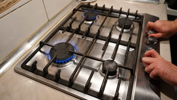 Turns off or turns on the gas on the stove. Blue flame of fire on black burners. The man's hands turn off or turn on the gas. Black grilles on a steel plate. White tiles on the walls. Cold winter