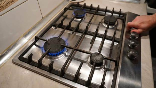 Turns off or turns on the gas on the stove. Blue flame of fire on black burners. The man's hand turns off or turns on the gas. Black grilles on a steel plate. White tiles on the walls. Cold winter