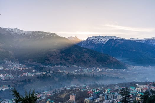 monsoon clouds moving over snow covered himalaya mountains with the blue orange sunset sunrise light with town of kullu manali valley at the base of mountains India