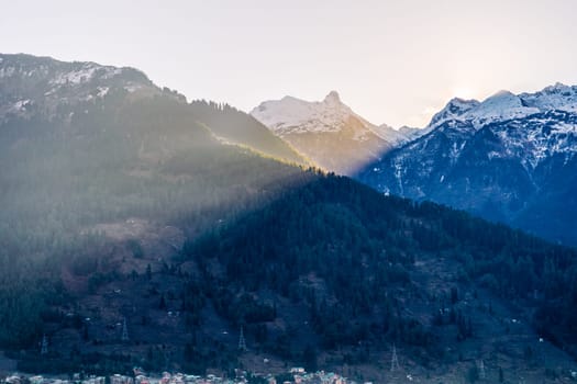 Light rays from sunrise behind himalaya mountains flooding light over half of tree covered mountains with manali town at base in India