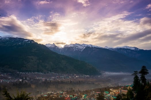 monsoon clouds moving over snow covered himalaya mountains with the blue orange sunset sunrise light with town of kullu manali valley at the base of mountains India