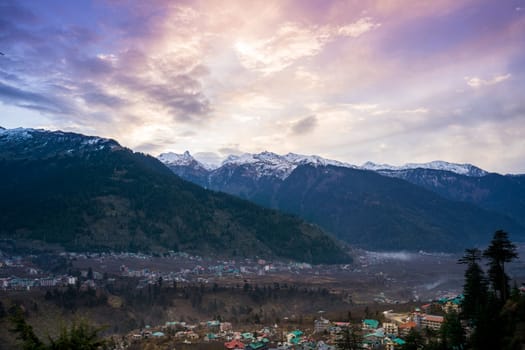 monsoon clouds moving over snow covered himalaya mountains with the blue orange sunset sunrise light with town of kullu manali valley at the base of mountains India