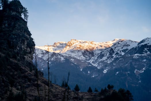 Sunlight lit peaks covered with snow in the distance with tree covered mountains in the foreground in manali India