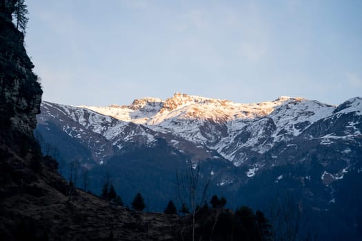 Sunlight lit peaks covered with snow in the distance with tree covered mountains in the foreground in manali India