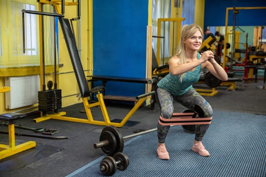 Using an exercise rubber band to enhance the effect of the exercise. A young girl dressed in leggings and a tight blue top exercises at the gym. The woman is performing squats. Blue walls can be seen in the background.