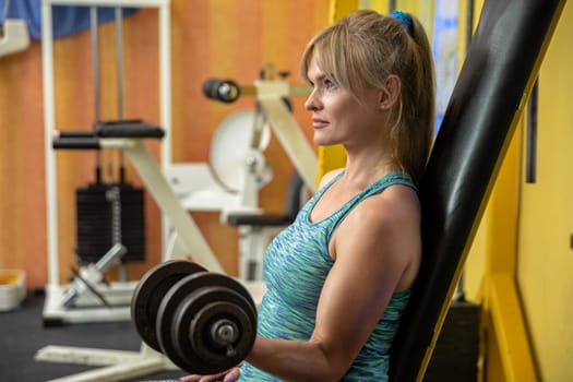 The woman holds a weight in her hand and lifts it, looking ahead. Profile view of a silhouette of a woman sitting on an exercise machine. A girl is lifting weights at the gym. Blurred background.
