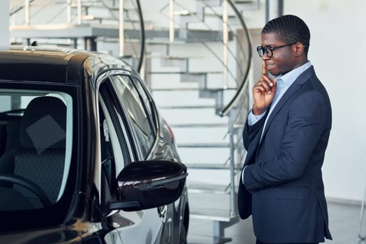 Standing near car. Young african american businessman in black suit is the autosalon.