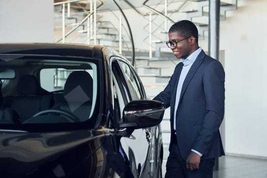 Standing near car. Young african american businessman in black suit is the autosalon.