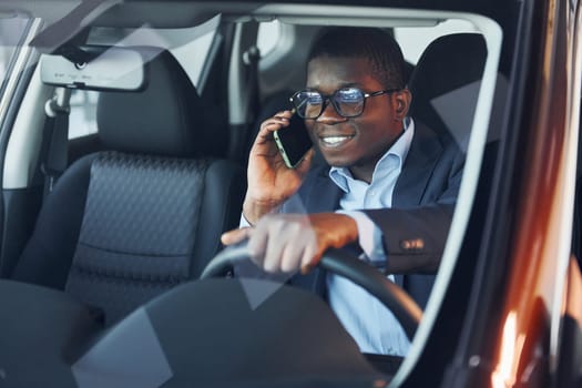 With phone. Young african american businessman in black suit is in the automobile.