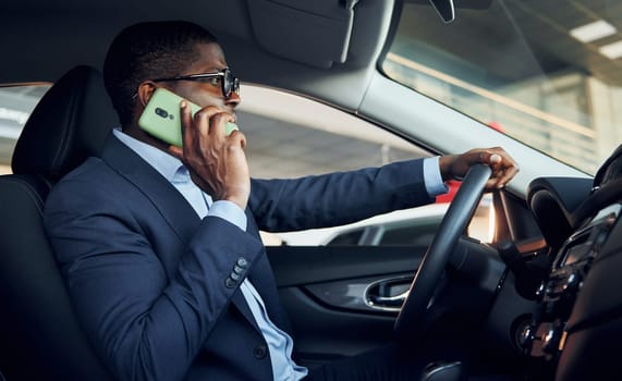 With phone. Young african american businessman in black suit is in the automobile.