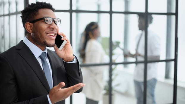 Close-up portrait of a smiling young african man talking on a mobile phone in the office against the background of colleagues