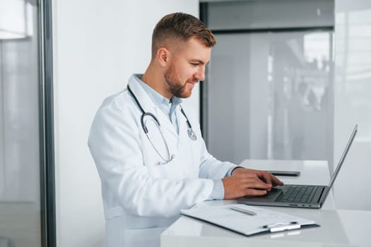 Using laptop. Professional medic in white coat is in the clinic.