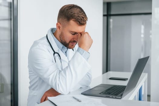 Using laptop and feeling tired. Professional medic in white coat is in the clinic.