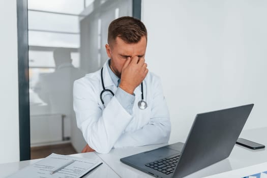 Using laptop and feeling tired. Professional medic in white coat is in the clinic.