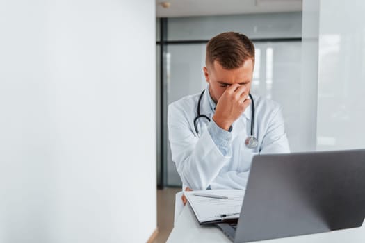 Using laptop and feeling tired. Professional medic in white coat is in the clinic.