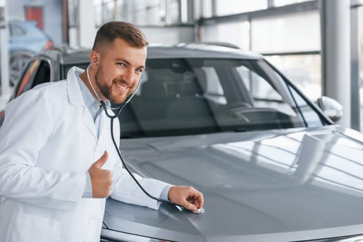 Showing thumb up. Medic in white coat is using stethoscope on the automobile.