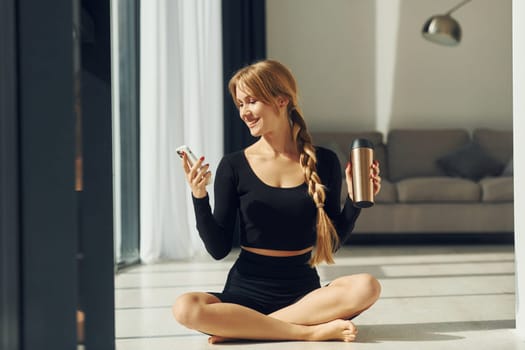 With phone in hands. Woman in sportive clothes doing yoga indoors.