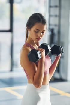 Holding dumbbells. Beautiful young woman with slim body type is in the gym.